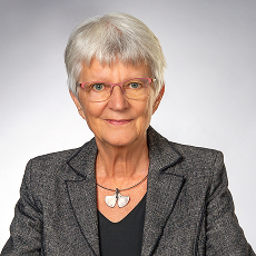 Eine ältere Frau mit kurzen grauen Haaren und Brille, die einen grauen Blazer, ein schwarzes Oberteil und eine Halskette mit zwei silbernen blattförmigen Anhängern trägt, posiert vor einem schlichten hellgrauen Hintergrund.