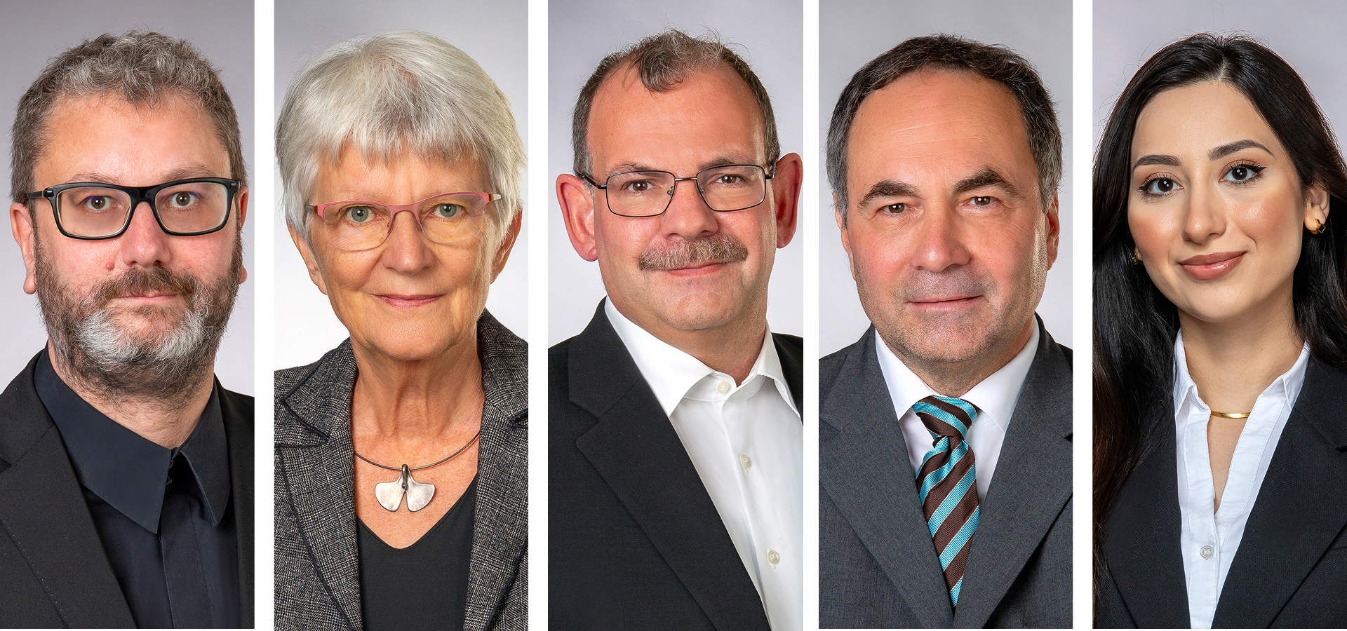 Eine Gruppe von fünf professionell gekleideten Personen, drei Männer und zwei Frauen, stehen nebeneinander vor einem neutralen Hintergrund, schauen in die Kamera und lächeln leicht.