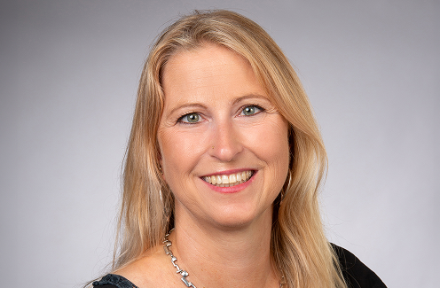 Lächelnde Frau mit langen blonden Haaren, die ein dunkles Oberteil und eine silberne Halskette trägt und vor einem schlichten hellgrauen Hintergrund posiert.