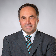 Ein Mann mittleren Alters im grauen Anzug, weißem Hemd und gestreifter Krawatte posiert für ein professionelles Portraitfoto vor einem schlichten hellgrauen Hintergrund. Er hat kurzes dunkles Haar und ein leichtes Lächeln.