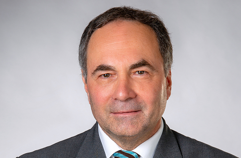 Mann mittleren Alters mit kurzen grauen Haaren, der einen dunklen Anzug, ein weißes Hemd und eine gestreifte Krawatte trägt und vor einem schlichten hellgrauen Hintergrund posiert.