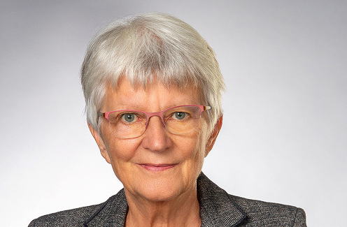Eine ältere Frau mit kurzen grauen Haaren, die eine rosafarbene Brille und einen grauen Blazer trägt, lächelt sanft in die Kamera vor einem schlichten hellen Hintergrund.