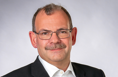 Mann mit kurzen Haaren, Brille und Schnurrbart, der eine dunkle Anzugsjacke und ein weißes Hemd trägt und vor einem schlichten hellen Hintergrund leicht lächelt.