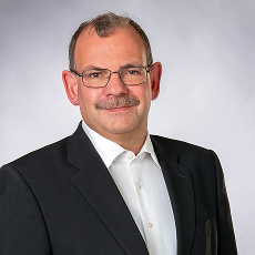 Ein Mann mittleren Alters mit kurzen Haaren, Brille und Schnurrbart trägt eine dunkle Anzugsjacke und ein weißes Hemd. Er lächelt leicht und posiert vor einem schlichten hellgrauen Hintergrund.