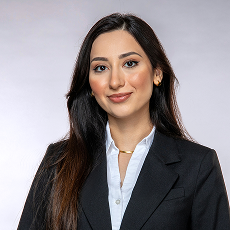 Eine Frau mit langen dunklen Haaren, die einen schwarzen Blazer, ein weißes Hemd und Goldschmuck trägt, lächelt sanft in die Kamera vor einem hellgrauen Hintergrund.