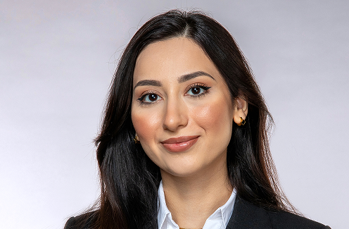 Eine Frau mit langen dunklen Haaren, die einen schwarzen Blazer, ein weißes Hemd und goldene Ohrringe trägt, lächelt sanft in einem professionellen Studio-Porträt vor einem hellgrauen Hintergrund.