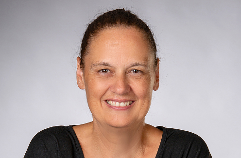 Eine Frau mittleren Alters mit zurückgebundenen braunen Haaren lächelt in die Kamera. Sie trägt ein schwarzes Oberteil und posiert vor einem schlichten, hellgrauen Hintergrund.