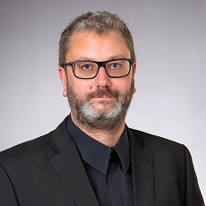 Ein Mann mit kurzen grauen Haaren, einem Bart und einer Brille trägt einen schwarzen Anzug und ein schwarzes Hemd und posiert vor einem schlichten hellgrauen Hintergrund.