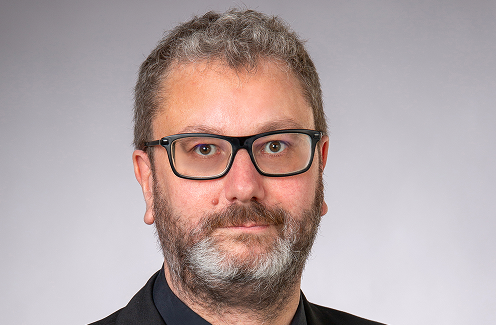 Ein Mann mit kurzem, lockigem, grauem Haar und Bart, der eine schwarze rechteckige Brille und eine dunkle Jacke trägt, blickt vor einem schlichten, hellgrauen Hintergrund direkt in die Kamera.